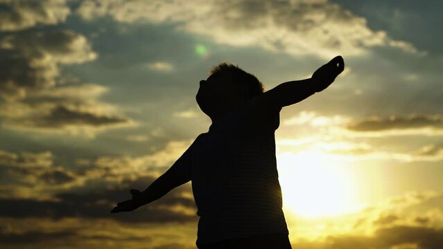 Child plays in park against sky. Little boy prays against background of sky and sun. Child raised his hands to sky in park at sunset, true faith. Religion and god, childhood dreams. Happy family
