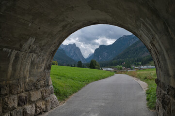 Obraz premium Blick durch eine gemauerte Unterführung auf einen Radweg der in Richtung eines Ortes mit Bergpanorama im Hintergrund läuft.
