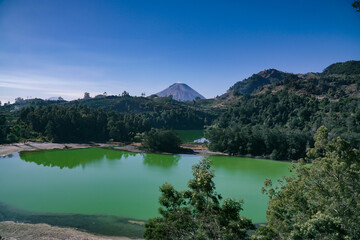 Green water lake surrounded by dense trees of forest and mountain on the background. Telaga Warna, Dieng Plateau. Indonesia