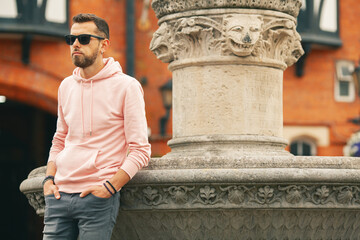 Travel concept. Young bearded romantic man wearing pink trendy hoodie, gray jeans posing over brick...