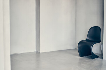 A plastic curved chair in black against a gray, rough, uneven wall with recesses and with space for...