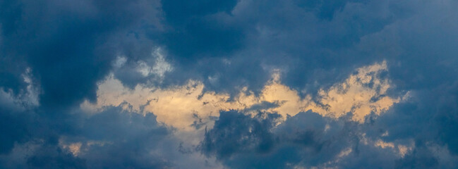Naklejka na ściany i meble A patch of sky illuminated by the evening sun shines through the dark blue clouds