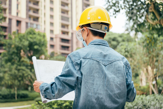 A middle-aged Chinese male engineer wearing a safety hard hat, a disposable mask and a pair protective goggles in a residential area is looking at the architectural drawing in his hands. 