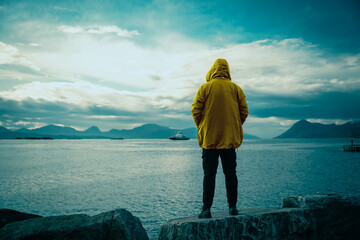 Guy standing on the rock and admiring a beautiful scenery of Norway fjord and mountains. Rainy weather and warm clothing.