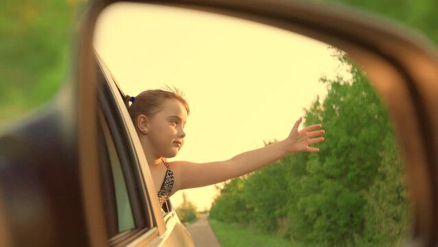 Child smiles, enjoys family vacation, car trip. Happy child is driving in car, looking out window with an outstretched hand. Little girl, child traveling by car, Video in rear view mirror. Child trip