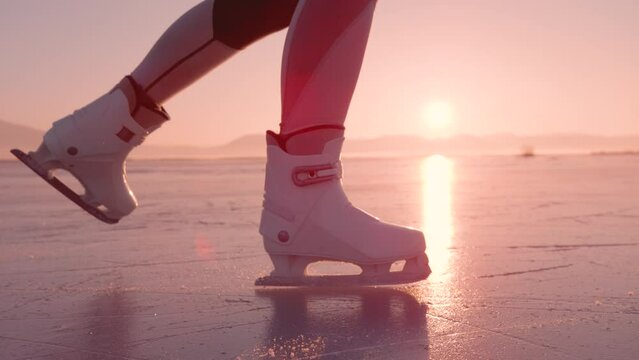 SUPER SLOW MOTION, CLOSE UP, LENS FLARE: Female skates on glittering ice surface of a frozen lake at sunrise. Woman is gliding on naturally frozen ice with her white skates on a sunny winter day.