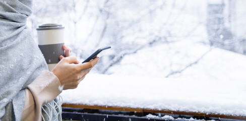 A woman in a warm hat and blanket holds smartphone in her hands. Winter landscape with snowfall in...