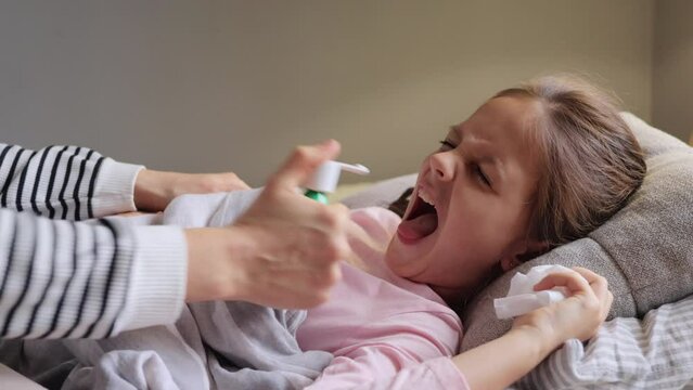 Domestic treatment. Childhood sickness. Kid's fever. Sick little girl has fever lying in bed having sore throat symptoms treating it with tonsil spray opening mouth wide while mother helps her.