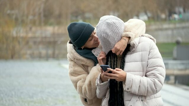 Happy dominican woman covering her girlfriend's eyes and surprising her while she is using her smartphone at street in winter.