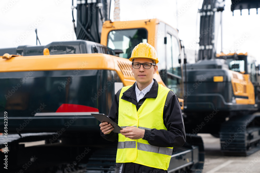 Aufkleber engineer in a helmet with a digital tablet stands next to construction excavators.