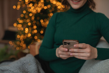 Close-up of a girl sitting with the phone at home near the decorated Christmas tree and sending congratulations to her friends. Holiday concept.