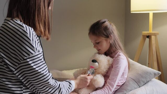 Mother and little daughter playing doctor at home while kid on sick leave using stethoscope to listening teddy bear having fun while child catching cold.