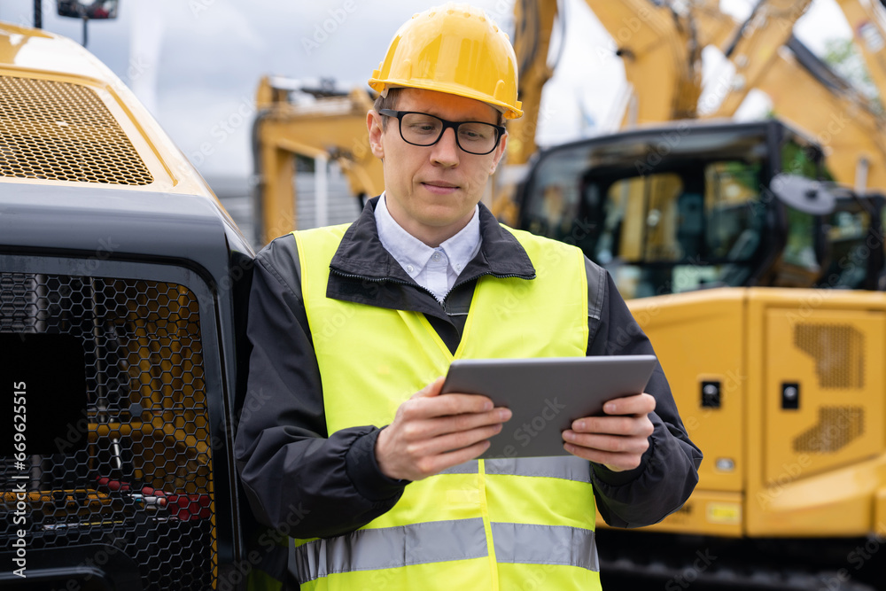 Aufkleber engineer in a helmet with a digital tablet stands next to construction excavators.