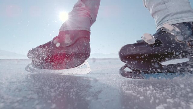 LENS FLARE, SUPER SLOW MOTION, CLOSE UP: Ice skater stops in front of camera. Unknown man is practicing a hockey stop while ice skating on a sparkling surface of a frozen lake on a sunny winter day.