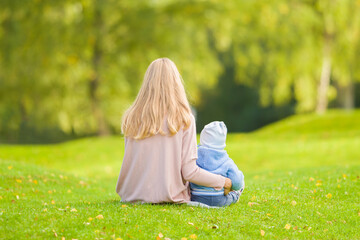 Fototapeta premium Young adult mother and baby boy sitting on green grass at park. Spending time together in beautiful autumn day. Back view. Lovely emotional moment. Peaceful atmosphere in nature. Closeup.