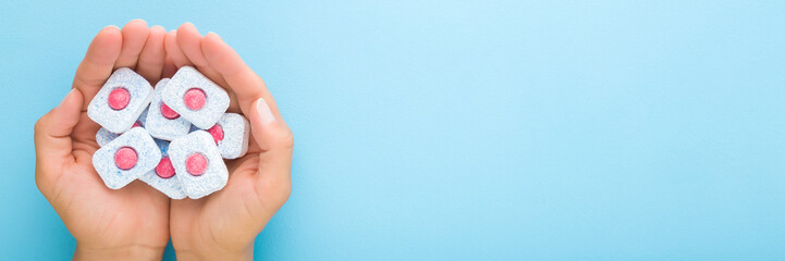 Young woman opened palms holding and showing red blue dishwasher tablets on light blue table background. Pastel color. Closeup. Point of view shot. Wide banner. Empty place for text. Top down view.