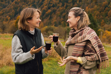 Two laughing women with mulled wine on autumn nature background. Two sisters having fun. Cheerful girls in autumn mountains. Happy friends in natural park. Weekend together with family.