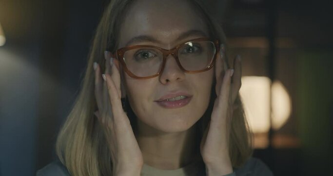 Close-up portrait of cheerful young lady with blond hair and eyeglasses smiling looking at camera in office at night. Businessperson and job concept.