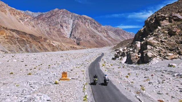 An aerial drone shot of male bike riders with luggage traveling on an empty scenic road in a cold mountainous region next to a river valley of Ladakh, India. Ladakh's beautiful mountain landscape.