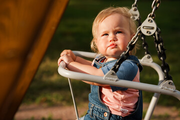 Baby kid in a denim jumpsuit swings on a swing in the summer in a public park, a place for text. The concept of a happy childhood. Active outdoors leisure for child.