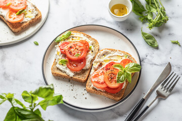 Sandwiches or toasts with tomatoes, cream cheese, olive oil and basil on a plate on white marble background. Traditional italian mediterranean food