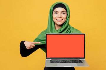 Naklejka premium Young asian muslim IT woman in green hijab abaya black clothes work use point on blank screen laptop pc computer isolated on plain yellow background People uae middle eastern islam religious concept