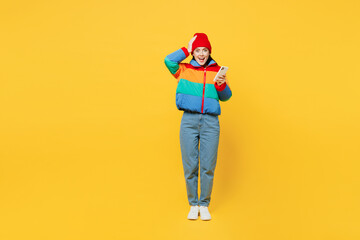 Full body young astonished cool happy woman she wearing padded windbreaker jacket red hat casual clothes hold head use mobile cell phone isolated on plain yellow background studio. Lifestyle concept.