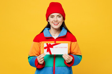 Young smiling happy woman she wears padded windbreaker jacket red hat casual clothes hold gift certificate coupon voucher card for store isolated on plain yellow background studio. Lifestyle concept. - obrazy, fototapety, plakaty