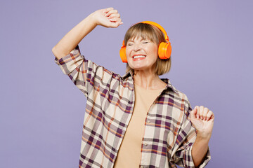 Elderly blonde fun woman 50s years old wear beige t-shirt shirt casual clothes listen to music in headphones raise up hands dancing isolated on plain pastel light purple background. Lifestyle concept.