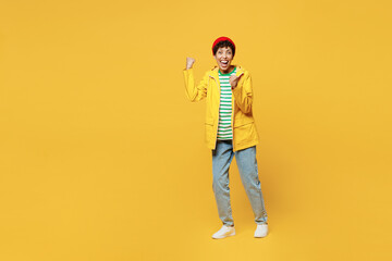 Full body young woman wear waterproof raincoat outerwear red hat do winner gesture celebrate clench fists isolated on plain yellow background studio. Outdoor lifestyle wet fall weather season concept