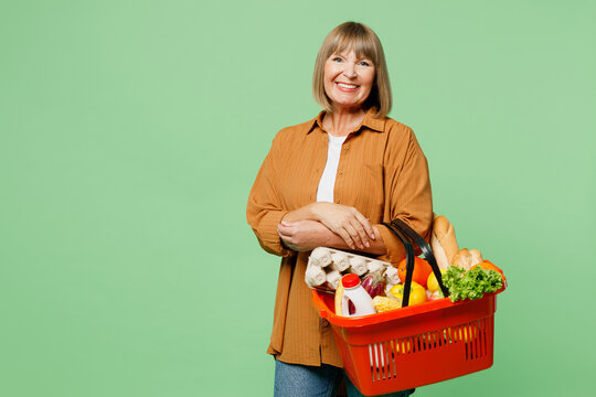 Side view elderly smiling happy cheerful woman wear brown shirt casual clothes hold shopping basket bag with food products look camera isolated on plain green background. Delivery service from shop.