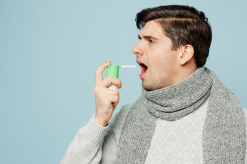 Young sad ill sick man wear gray sweater scarf use throat spray against cough isolated on plain blue background studio portrait. Healthy lifestyle disease virus treatment cold season recovery concept.