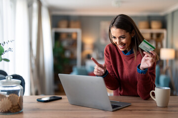 Happy young businesswoman holding credit card using online bank app on laptop making convenient...