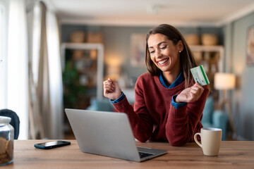 Happy young female shopper holding credit card, using laptop doing online banking transaction while sitting at desk at home. - obrazy, fototapety, plakaty