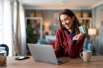 Smiling young adult woman holding credit card while shopping online over laptop computer.