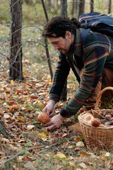 Young male backpacker bending over boletus and cutting it by knife while picking mushrooms in autumn forest on weekend