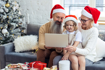 Concept of spending vacations or holidays at grandparents. Attractive active grandfather and grandmother, senior man and woman, with grandchild doing video call with parents on laptop or smartphone