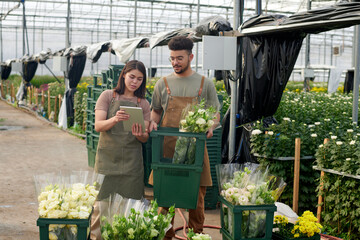 Young confident female clerk of flowers showing her colleague new sorts on tablet screen while both standing in spacious industrial greenhouse
