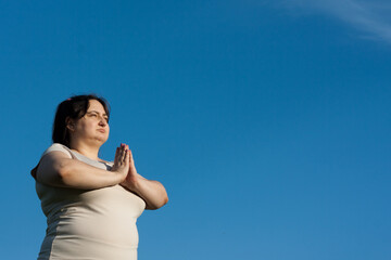 Portrait of middle-aged woman against blue sky. Calm woman holding hands in namaste mudra during meditation. Overweight woman practices yoga outdoors in summer. Mental health, healthy lifestyle