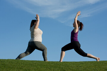 Two middle-aged women practice yoga in city park in warrior pose, blue sky background. Healthy...