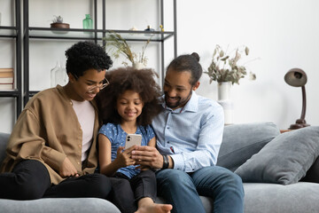 Happy young hipster Black parents and pre teen gen Z kid using online learning app on cellphone,...