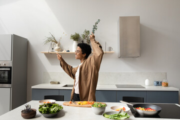 Excited active Black cooking blogger woman having fun in home kitchen, dancing to music at table...
