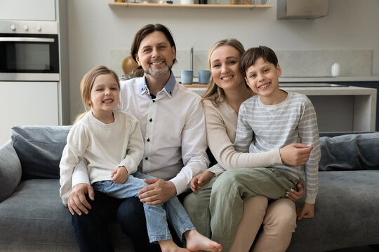 Joyful parents holding little daughter and teenage son kid on laps, looking at camera, smiling, sitting on sofa in living room. Happy family couple and children at comfortable home.