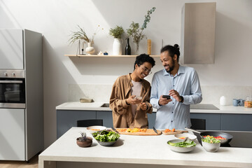 Joyful dating young African couple using online app on smartphones, cooking together in home...