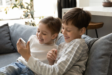 Excites cheerful gen Z little children using smartphone together, playing virtual video game, watching funny movie, talking on video call. Teen brother showing little sister app on cellphone