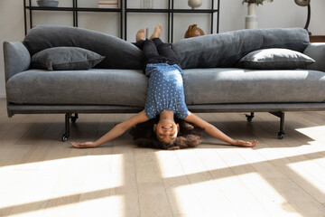 Excited funny cute Black girl kid with curly hair resting on comfortable sofa upside down with head on floor, smiling, laughing, playing active games, stretching body, enjoying being at cozy home