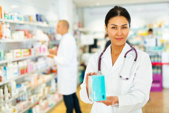 Oriental woman in lab coat standing in salesroom and holding medicine in hands. Her colleague working in background.