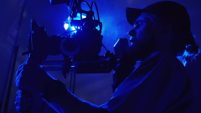 Medium close-up low-angle shot of young male videographer with professional camera on shoulder support with stabilizer working on commercial shoot, looking at viewfinder and giving instructions