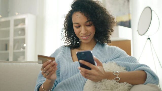 Smiling multi racial woman holding smartphone and credit card sitting on couch modern home. Happy diverse female shopper using instant easy mobile payments, customer making purchase online banking app