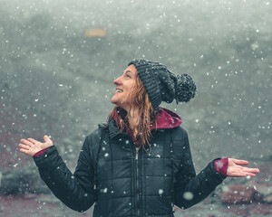 mulher com touca de lã e mãos levantadas, sorrindo, em cenário de inverno com neve caindo 